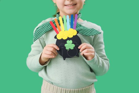 Cute little girl holding paper pot with coins and rainbow on green background. St. Patrick's Day celebration