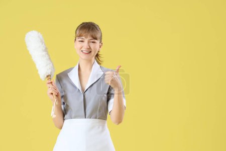 Junges Zimmermädchen mit Staubwedel zeigt Daumen nach oben auf gelbem Hintergrund