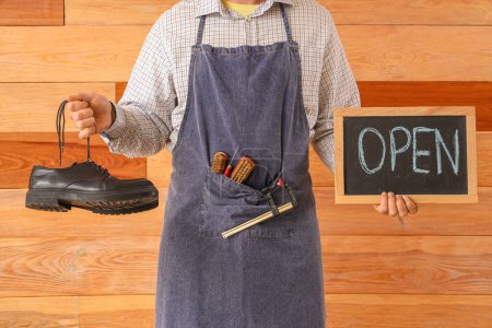 Male shoemaker with OPEN sign and boots on wooden background, closeup