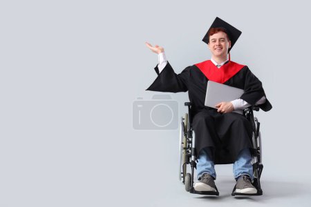 Männlicher Absolvent im Rollstuhl mit Laptop, der etwas auf hellem Hintergrund zeigt