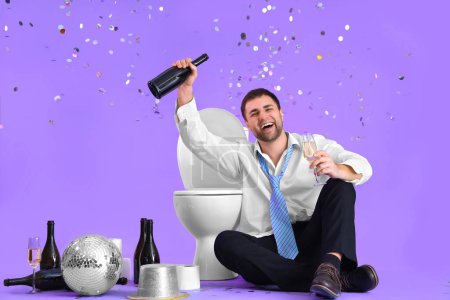 Téléchargez les photos : Jeune homme d'affaires ivre assis près de la cuvette des toilettes pendant la fête sur fond lilas - en image libre de droit