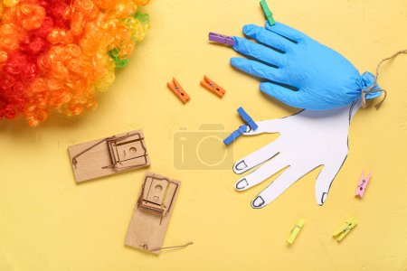 Medical glove with clothespins, clown wig and mousetraps on yellow background. April Fool Day