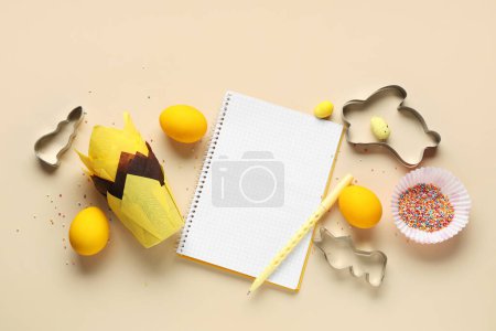 Téléchargez les photos : Bloc-notes vierge pour recettes avec stylo, oeufs de Pâques et outils de cuisson sur fond beige - en image libre de droit