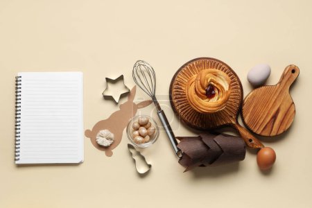 Téléchargez les photos : Bloc-notes vierge pour recettes avec pain sucré, décor de Pâques et outils de cuisine sur fond beige - en image libre de droit