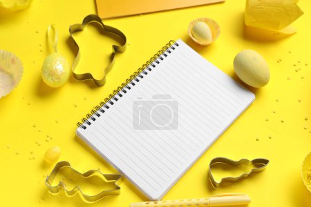 Téléchargez les photos : Bloc-notes vierge pour recettes avec stylo, oeufs de Pâques et outils de cuisson sur fond jaune - en image libre de droit