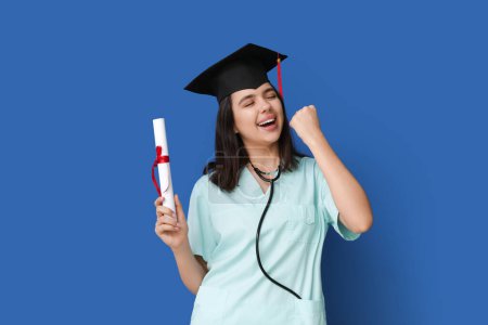 Photo for Happy female medical student in graduation hat with diploma on blue background - Royalty Free Image