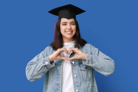 Photo for Happy smiling female student in graduation hat making heart with her hands on blue background - Royalty Free Image