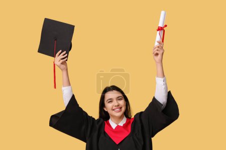Photo for Happy female graduating student holding diploma and graduation hat on yellow background - Royalty Free Image