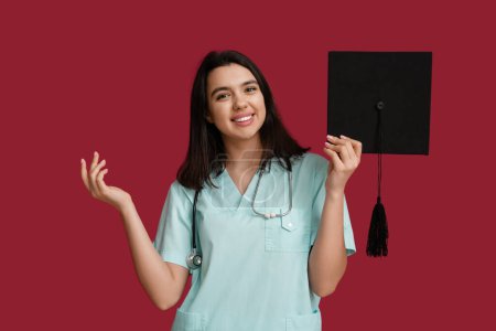 Photo for Happy female medical student holding graduation hat on red background - Royalty Free Image