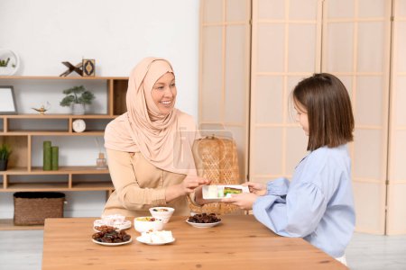 Glückliche muslimische Mutter und ihre Tochter sitzen am Tisch mit traditionellen Süßigkeiten im Wohnzimmer. Eid al-Fitr-Feier