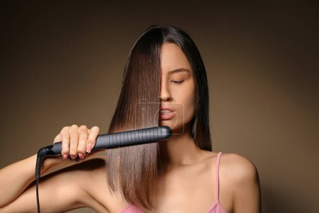 Beautiful young woman straightening hair on beige background