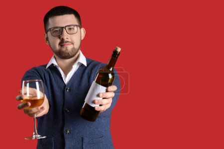 Young sommelier with glass and bottle of wine on red background