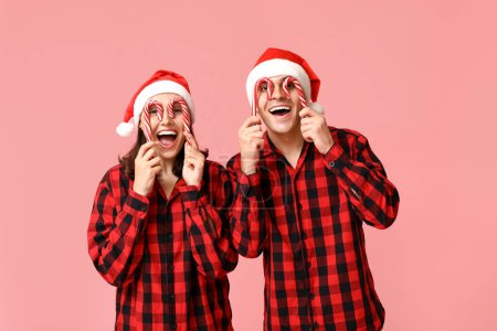 Foto de Pareja joven divertida en pijama de Navidad y con bastones de caramelo sobre fondo rosa - Imagen libre de derechos