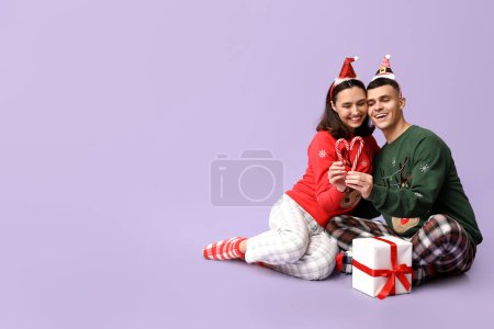 Foto de Feliz pareja joven en pijama de Navidad, con bastones de regalo y dulces sobre fondo lila - Imagen libre de derechos