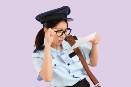 Junge asiatische Postbotin mit schlechter Sehkraft liest Adresse auf Umschlag vor fliederfarbenem Hintergrund