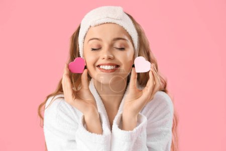 Beautiful young woman in bath headband with heart-shaped sponges on pink background, closeup