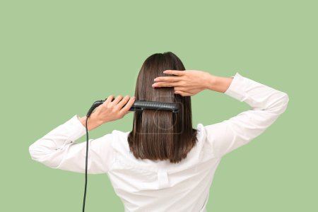 Photo for Beautiful young woman straightening hair on green background, back view - Royalty Free Image
