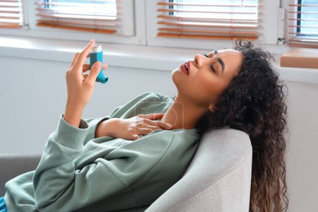 Téléchargez les photos : Jeune femme afro-américaine avec inhalateur ayant une crise d'asthme dans un fauteuil à la maison - en image libre de droit