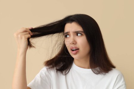 Stressed young woman with dandruff problem examining her hair on beige background