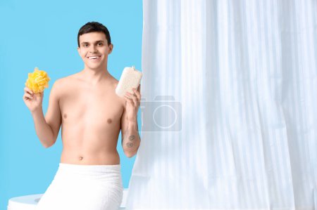 Young man with loofahs sitting on bathtub against blue background