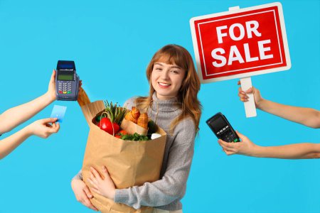 Young woman with shopping bag, payment terminals, credit card and FOR SALE sign on blue background
