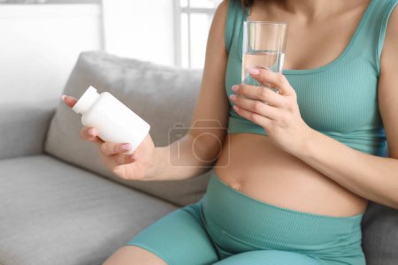 Photo for Sporty pregnant woman with pill bottle and glass of water sitting on sofa at home, closeup - Royalty Free Image