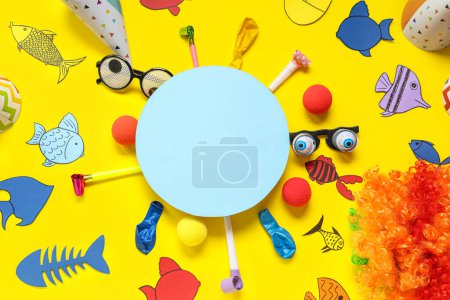 Blank card with paper fishes, clown wig and party decor on yellow background. April Fools Day celebration