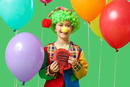 Funny little boy in clown costume with whoopee cushion and balloons on green background. April Fools Day celebration