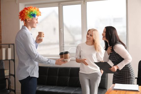Foto de Un joven con peluca graciosa dando café a sus colegas en la oficina. Celebración del Día de los Inocentes - Imagen libre de derechos