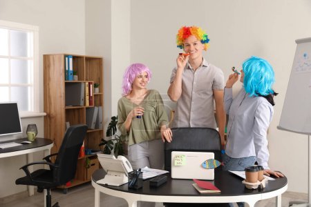 Foto de Gente de negocios con pelucas divertidas y silbatos de fiesta en la oficina. Celebración del Día de los Inocentes - Imagen libre de derechos