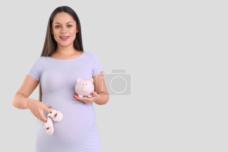 Young pregnant woman with piggy bank and baby booties on light background. Maternity Benefit concept
