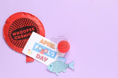 Festliche Postkarte zum Aprilscherz mit Kissen, Papierfischen und Clownsnase auf fliederfarbenem Hintergrund