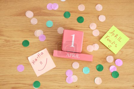 Würfelkalender mit Datum 1 APRIL, lustigen Papieraufklebern und Konfetti auf Holzgrund. Feier zum Aprilscherz