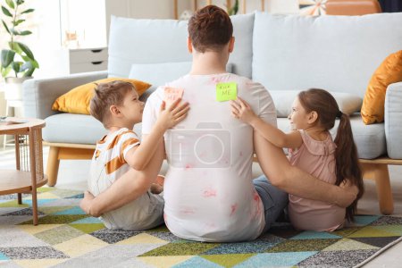 Kleine Kinder kleben ihrem Vater zu Hause KICK ME-Aufkleber auf den Rücken. Aprilscherz
