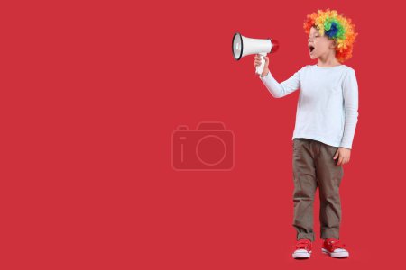Foto de Lindo niño en peluca de payaso colorido con megáfono sobre fondo rojo. Celebración del Día de los Inocentes - Imagen libre de derechos