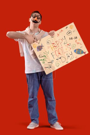 Foto de Joven disfrazado de gracioso apuntando al cartel con texto FELIZ DÍA DEL Tonto sobre fondo rojo - Imagen libre de derechos