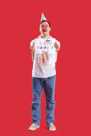 Foto de Joven con sombrero de fiesta y nariz de payaso sosteniendo cartel con texto 1 DÍA DE LOS TONOS DE ABRIL sobre fondo rojo - Imagen libre de derechos