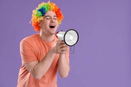 Foto de Hombre joven en peluca de payaso colorido con megáfono sobre fondo lila. Celebración del Día de los Inocentes - Imagen libre de derechos