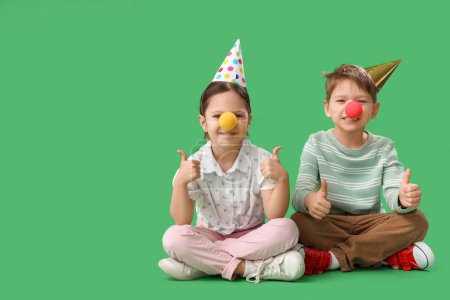 Little brother and sister with clown noses and party hats showing thumbs-up on green background. April Fool's Day celebration