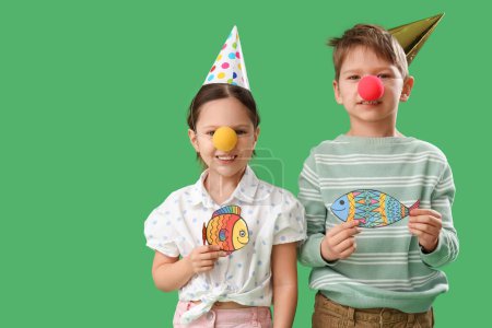 Foto de Hermano pequeño y hermana con narices de payaso, sombreros de fiesta y peces de papel sobre fondo verde. Celebración del Día de los Inocentes - Imagen libre de derechos