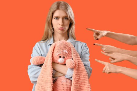People pointing at upset young woman with toy on orange background. Accusation concept