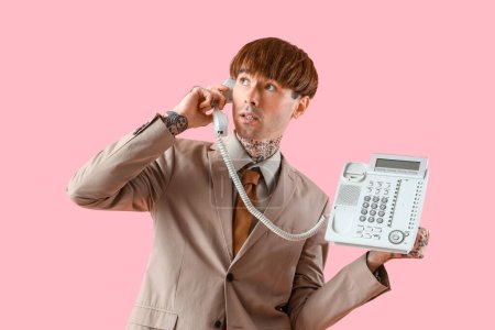 Handsome young tattooed businessman talking by landline phone on pink background