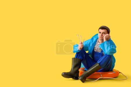 Photo for Upset young sailor in raincoat with wrenches sitting on ring buoy against yellow background - Royalty Free Image