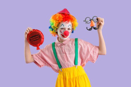 Funny little girl dressed as clown with disguise and whoopee cushion on lilac background. April Fools' Day celebration