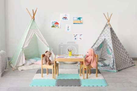 Photo for Cute toy rabbit and bear sitting at table near play tents in children's room - Royalty Free Image