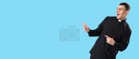 Happy young priest pointing at something on light blue background with space for text 