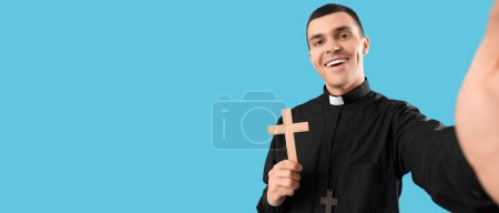 Young priest with cross taking selfie on light blue background with space for text 
