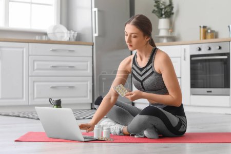 Mujer joven deportiva con ampolla de suplementos vitamínicos utilizando el ordenador portátil en la cocina