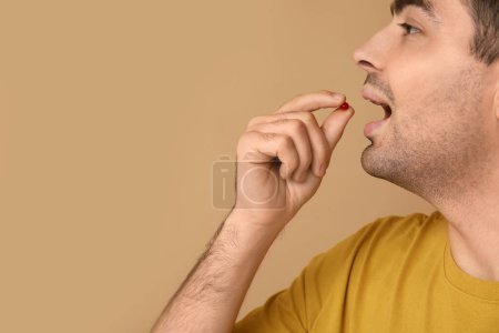 Young man taking vitamin A pill on beige background, closeup