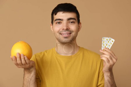 Young man with blisters of vitamin A pills and grapefruit on beige background